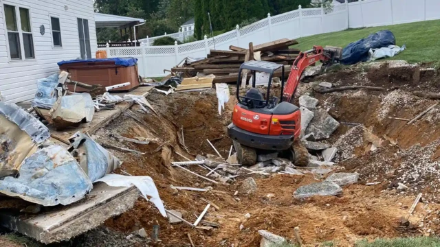 In the photo, you can see that we've successfully removed the fence and decking. We're currently placing the concrete outside the pit so we can load it into the dumptruck. A large construction pit filled with broken pool debris and concrete rubble, with a red Kubota excavator sitting inside. The destroyed pool is clearly visible with torn pieces of liner and scattered rebar.