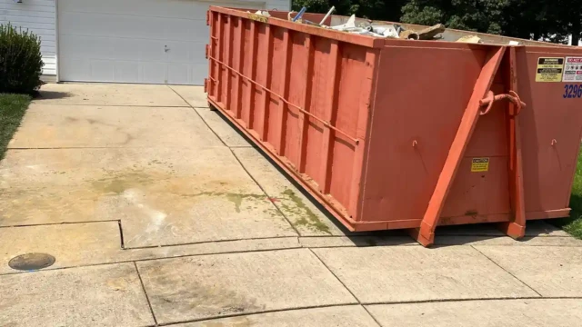 You can see our dumptruck full of rubble and debris, ready for disposal. A large orange dumpster sits on a driveway, filled with debris from the pool demolition. Some water stains and dirt are visible around the base on the concrete.